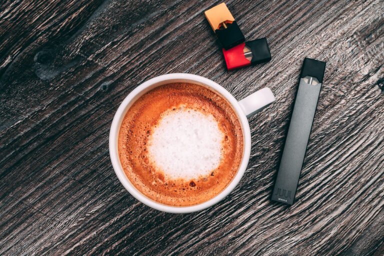Cup of cappuccino next to a pod vape and pods on a wooden table